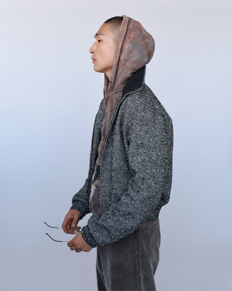 Asian man in profile wearing hoodie and tweed jacket stands indoors against a solid white backdrop looking pensive and stylish.