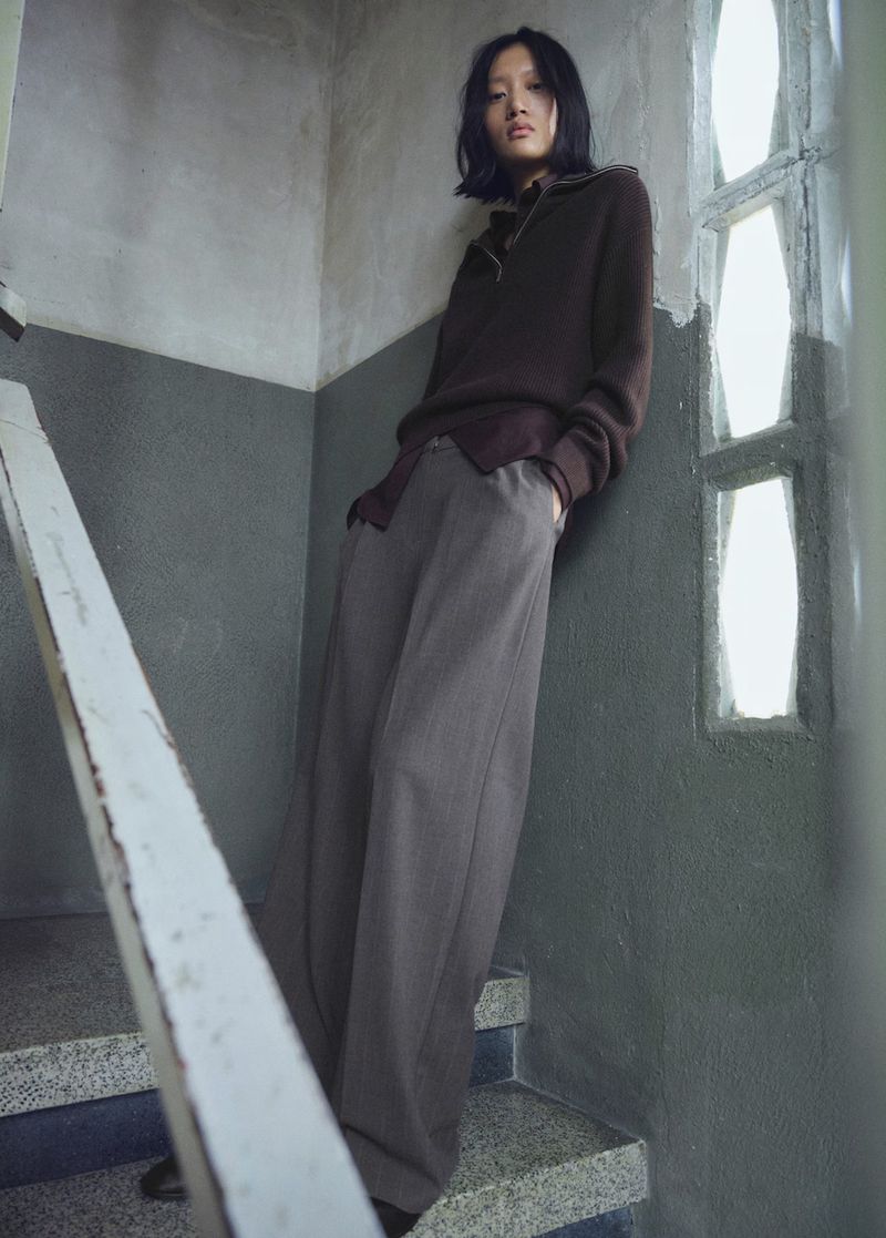 Fashion shot of a slender Asian woman in a brown sweater and wide grey pants posing on a staircase with a concrete wall and window.