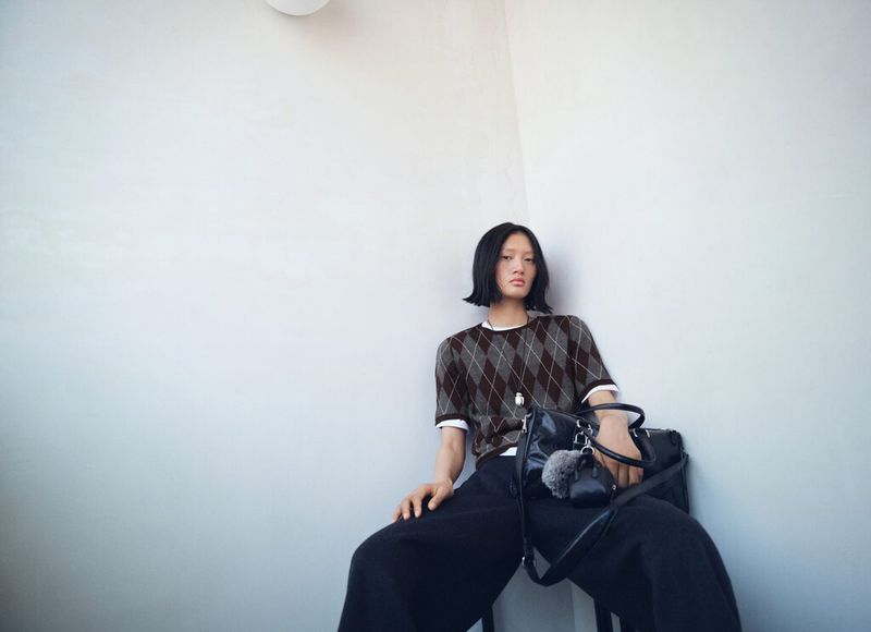 A model wearing a patterned brown top and wide-leg black pants sits against a white wall with a black bag, low-angle shot.
