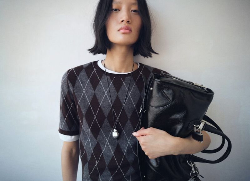Woman in argyle sweater holding a black handbag against a white wall, looking at the camera with a neutral expression.