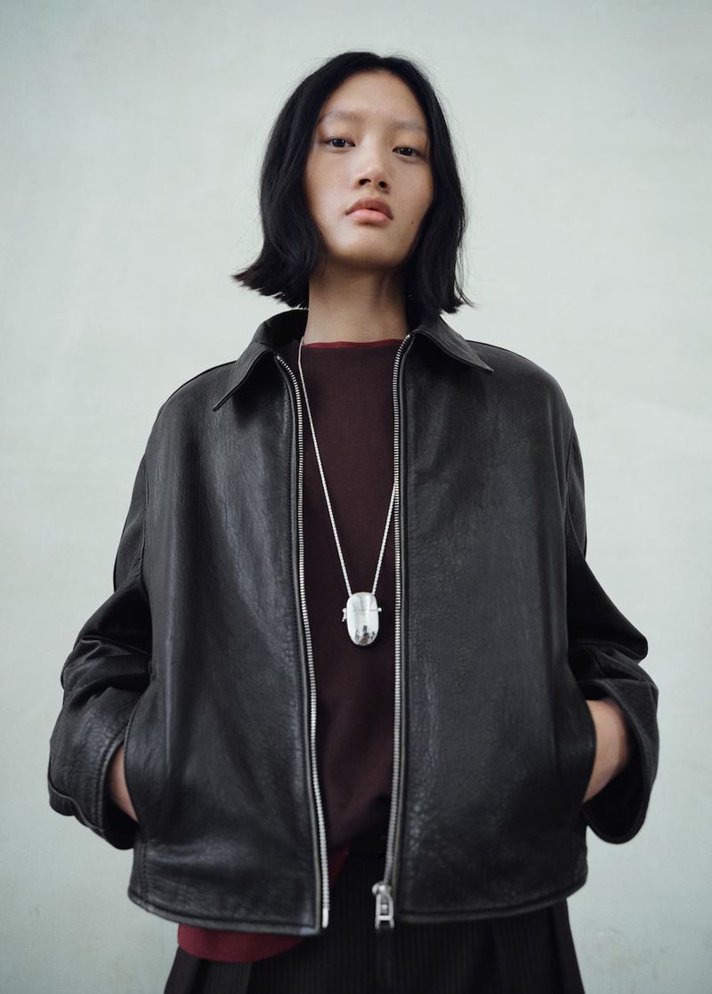 An Asian model wearing a black leather jacket and a silver pendant poses in a studio setting with a neutral background, capturing a fashion-forward look.