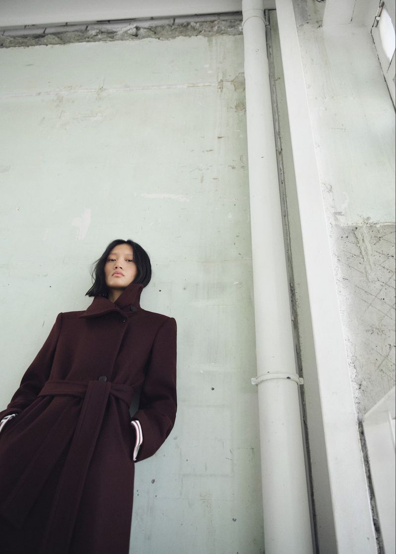 Fashion image of an Asian adult female model wearing a maroon coat with a tie belt, posing against a textured concrete wall indoors.