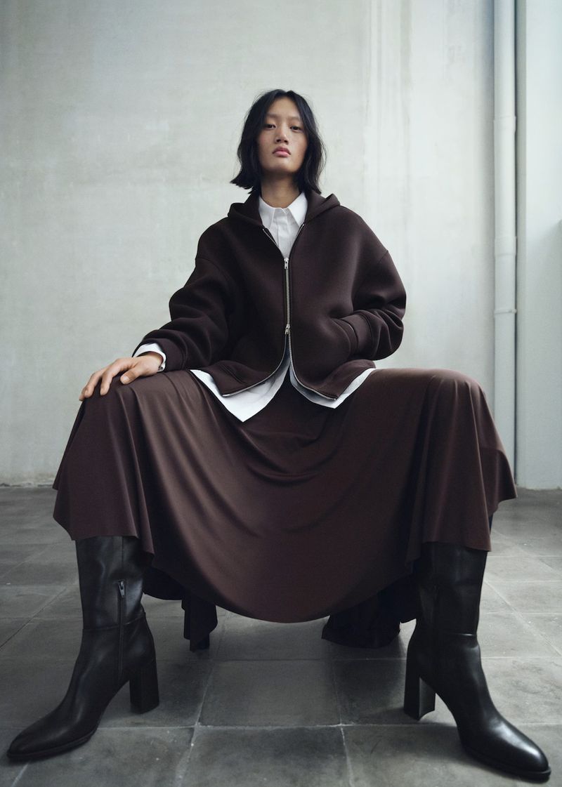 Asian woman poses in a brown hoodie, skirt, and black boots against a neutral studio backdrop, in a fashion editorial-style photograph.
