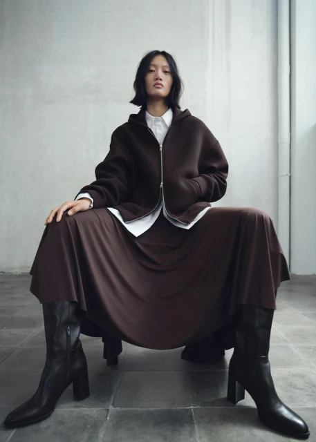 Asian woman poses in a brown hoodie, skirt, and black boots against a neutral studio backdrop, in a fashion editorial-style photograph.