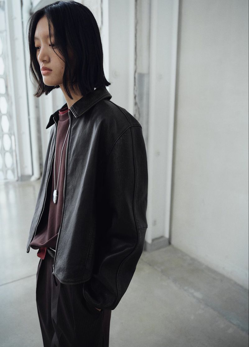 Adult female wearing a black leather jacket and burgundy top, standing indoors with a neutral expression in a simple setting.