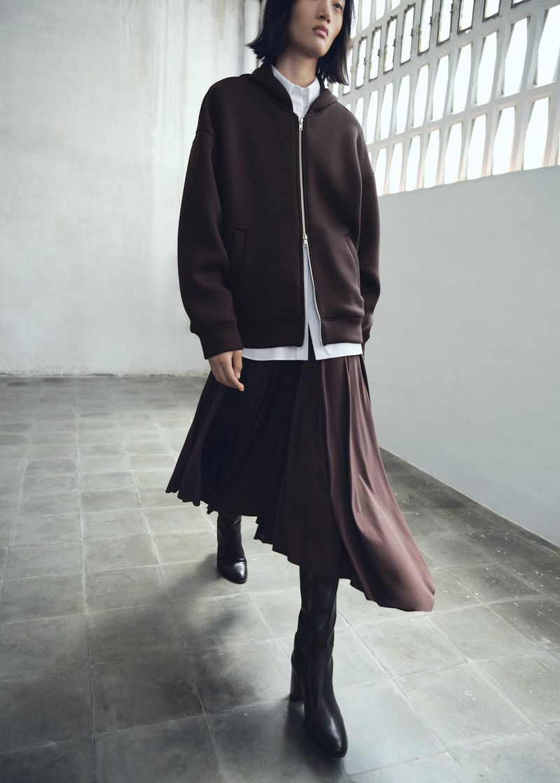 Fashionable adult wearing a brown zip-up jacket, white shirt, brown skirt, and black boots walking in a minimalist architectural setting.