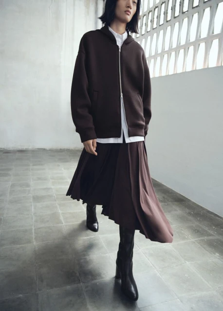 Fashionable adult wearing a brown zip-up jacket, white shirt, brown skirt, and black boots walking in a minimalist architectural setting.