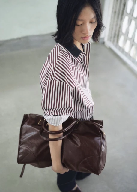 Asian woman in striped shirt carrying large brown leather duffel bag indoors with soft light and minimalistic style.
