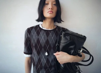 Model Yilan Hua wearing argyle sweater and holding black handbag against plain white background for Mango project Strictwear Burgundy