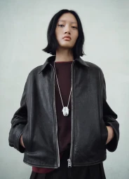 Model Yilan Hua wearing a dark leather jacket and burgundy sweater with a silver pendant necklace against a plain grey backdrop.