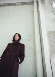 Model Yilan Hua posing in a tailored burgundy belted coat against a pale industrial wall with vertical pipes.
