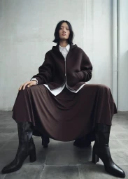 Model Yilan Hua seated wearing a dark brown hooded jacket, white shirt, skirt, and boots against a textured wall for Mango.