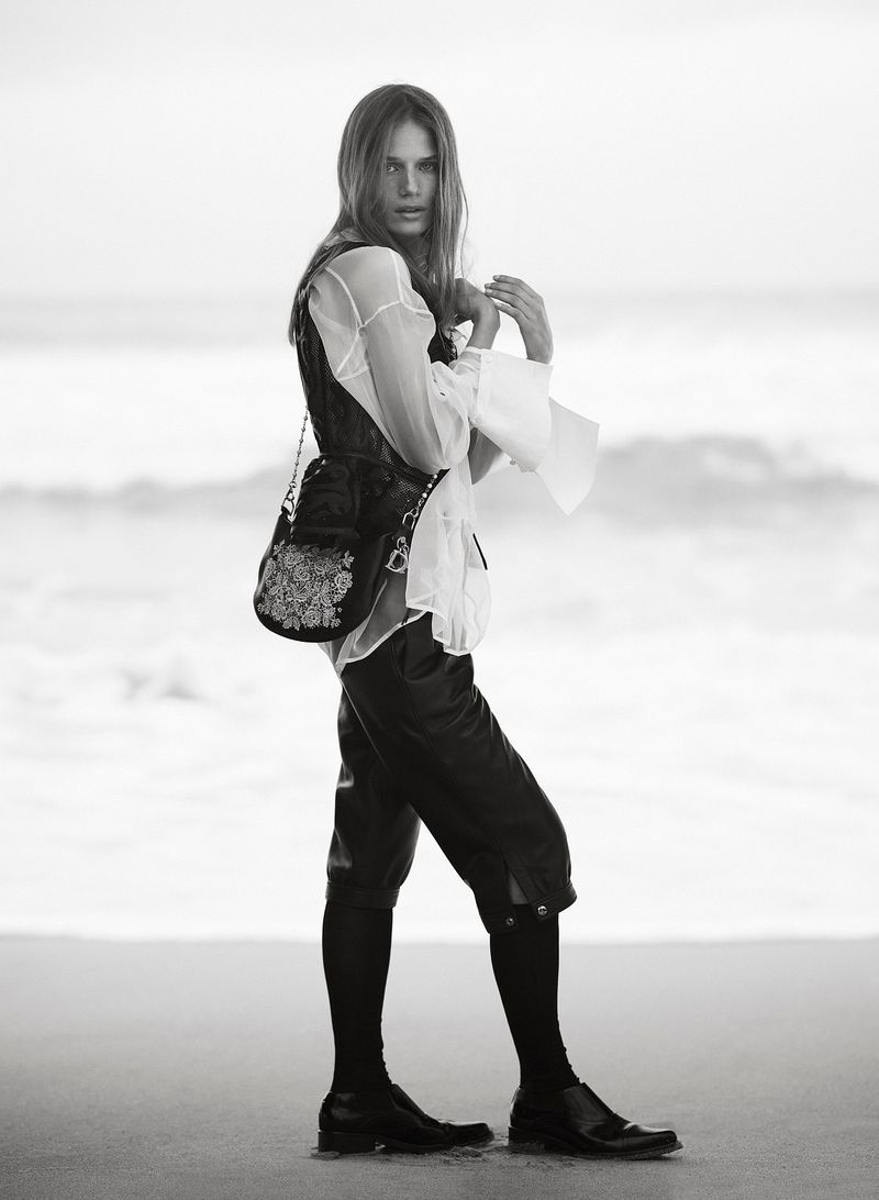 Monochrome image of a female model wearing a pirate-style outfit with a beaded bag on a beach, with soft waves in the background.