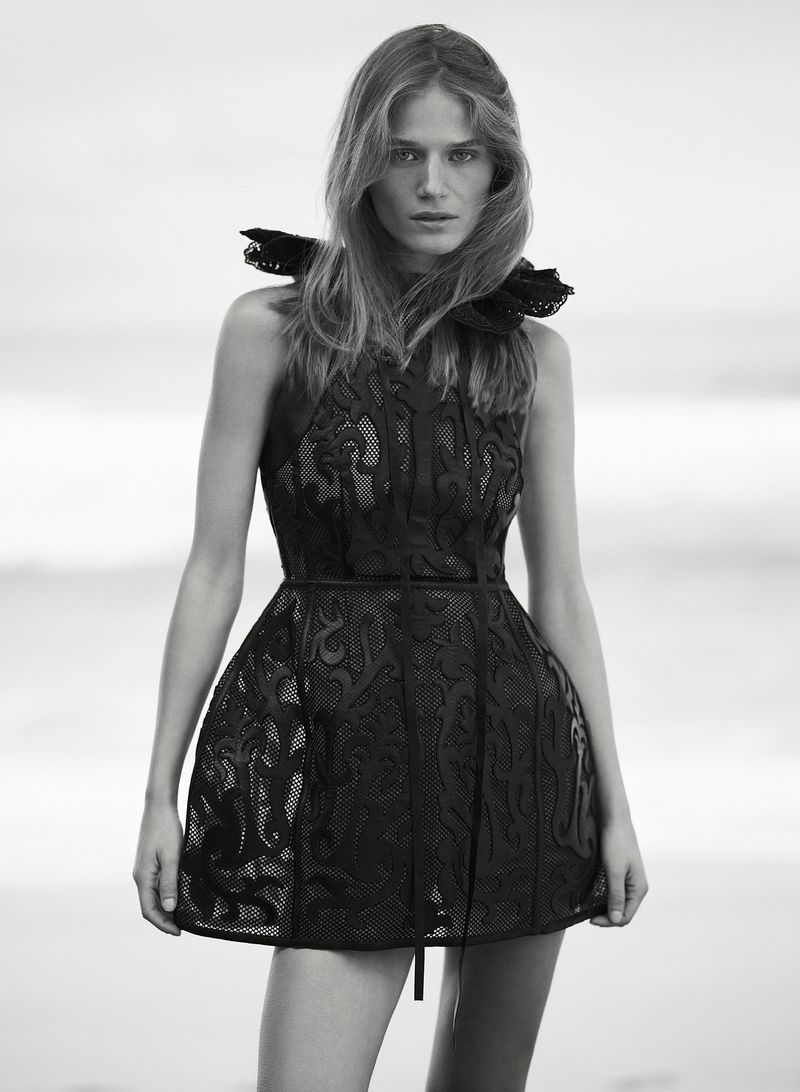 Black and white portrait of a fair-skinned young adult woman wearing a dark short dress with a frilled collar against a neutral background.