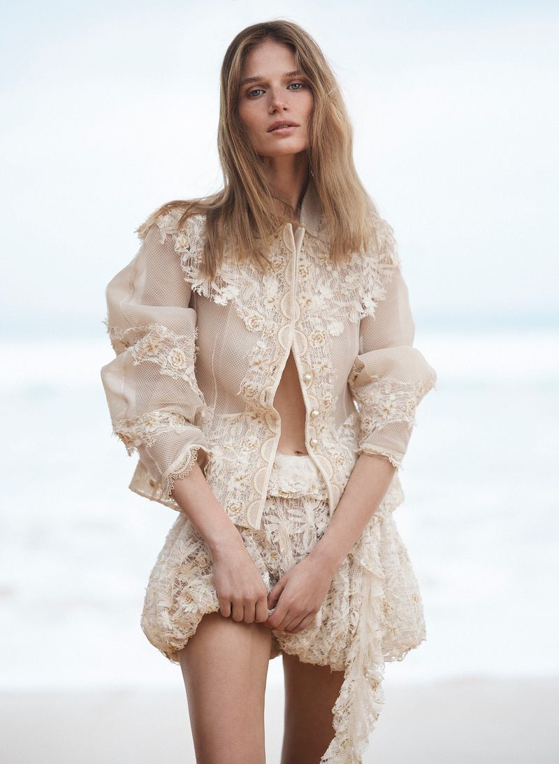 Fashion shot of a fair-skinned female model posing outdoors on a beach with a cloudy sky background, wearing an ornate lace blouse and shorts.