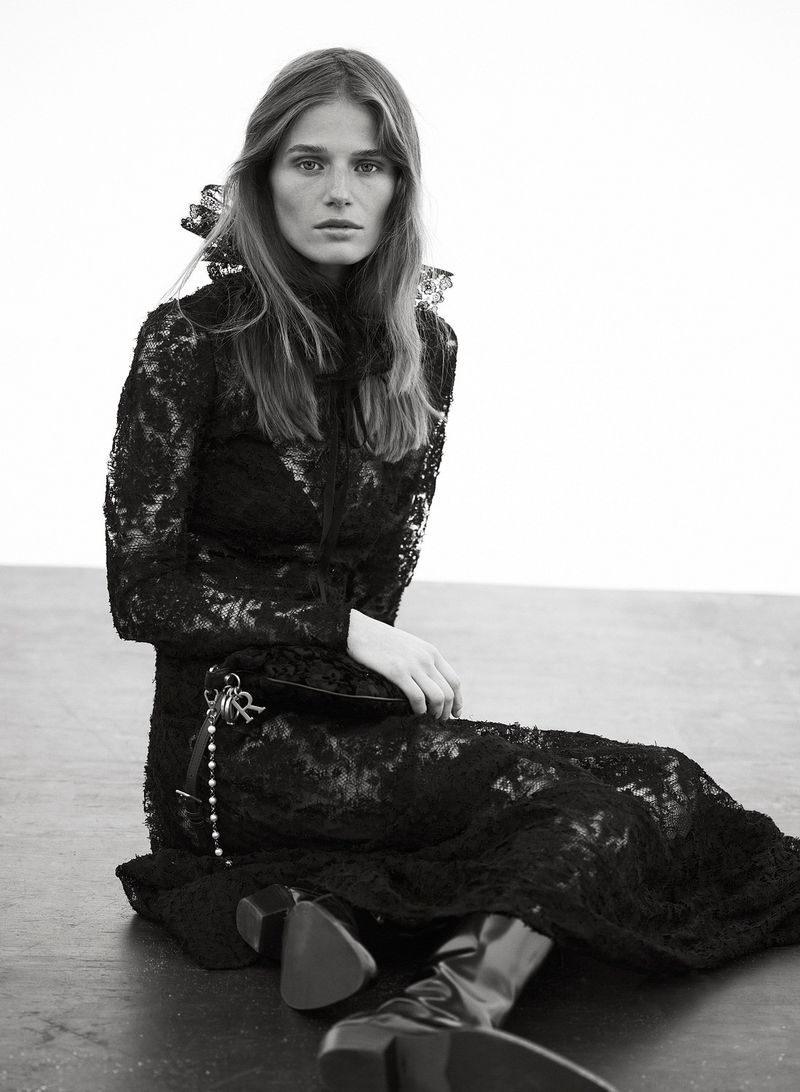 Monochrome fashion shot of a female model sitting in a black lace dress and black heeled boots against a white backdrop, studio setting.