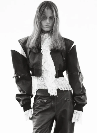 A slender model poses in an outdoor fashion shot, wearing a black leather jacket and pants with a frilly white lace blouse.