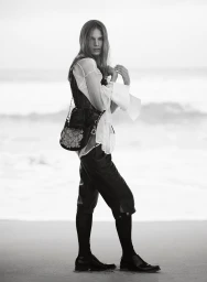 Model standing on a sandy beach wearing a sheer white blouse and dark trousers with a shoulder bag.