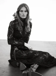 Model seated on the floor wearing a long-sleeved black lace dress with a ruffled collar and boots against a white wall.