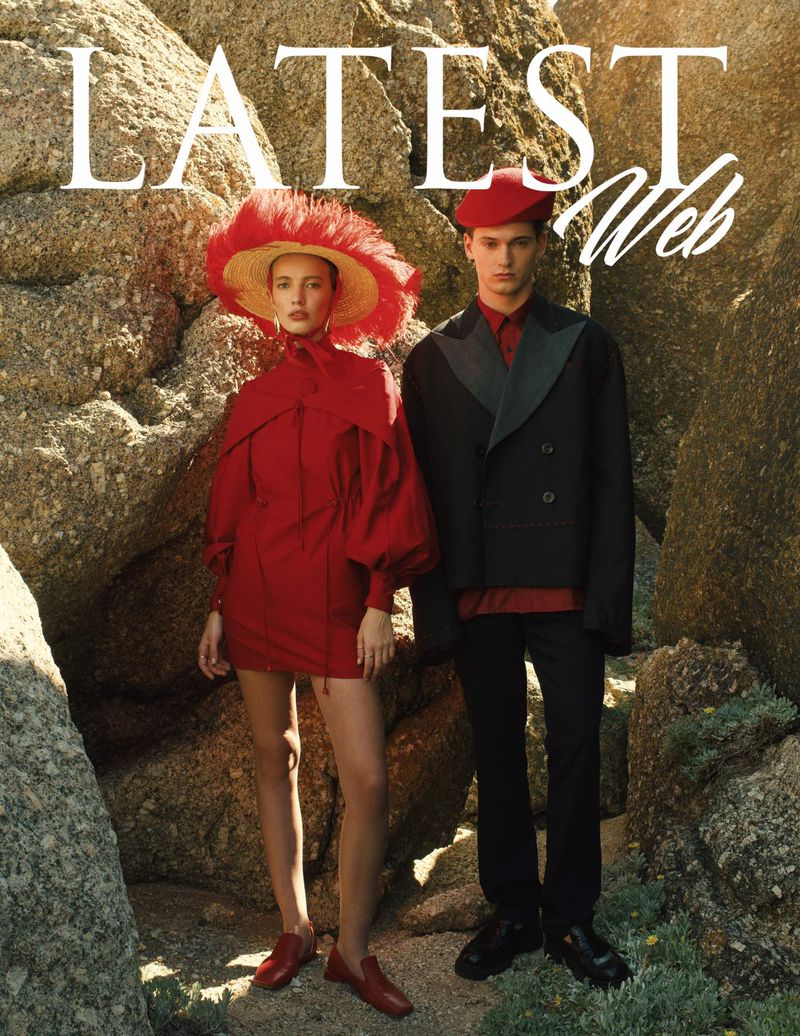 Editorial fashion shot of a young female model in red dress with wide hat standing next to a young male model in a blazer on rocky terrain.