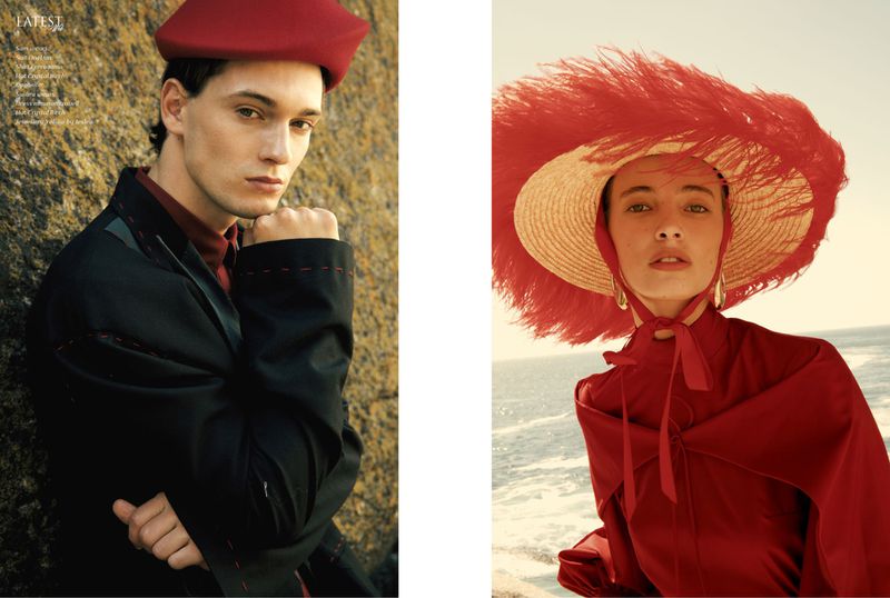 Fashion editorial featuring two adult models wearing matching red hats and formalwear, posing on a rocky seaside landscape at daytime.