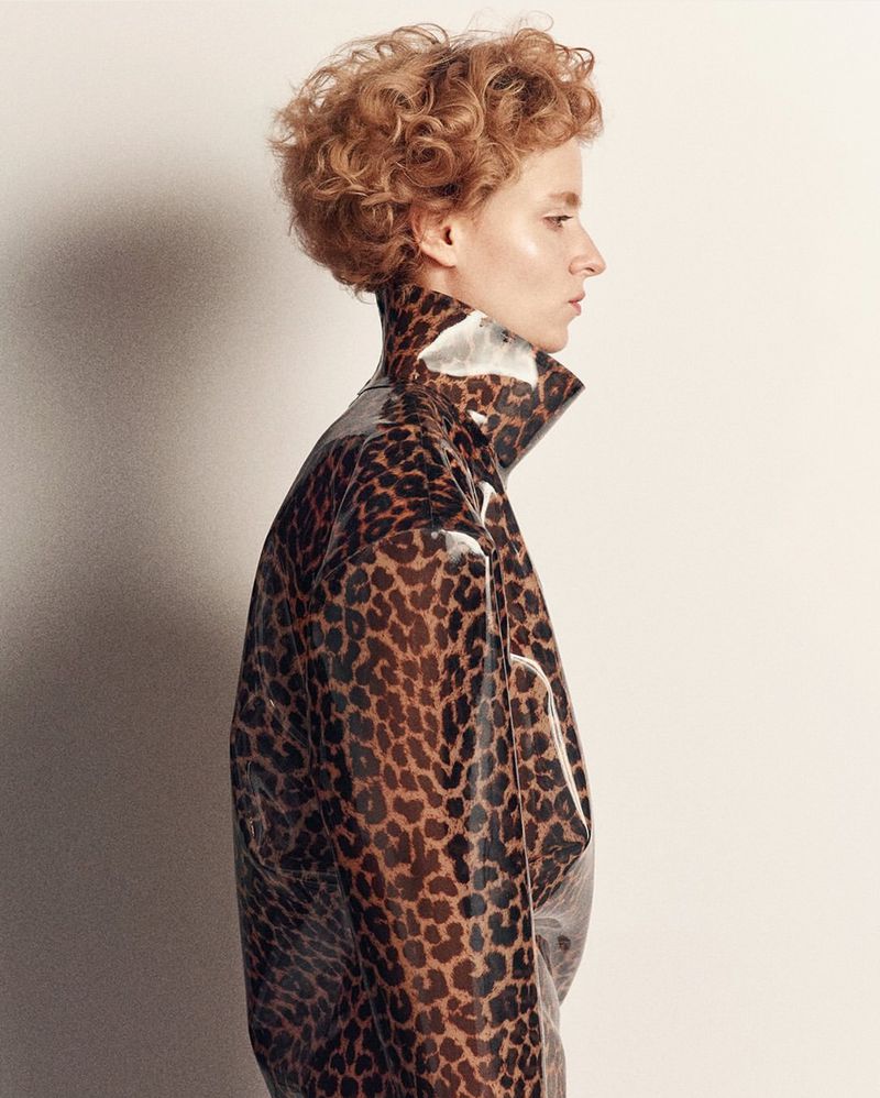 A woman with curly red hair wearing a shiny leopard-print jacket stands against a white backdrop, showcasing a stylish fashion portrait.