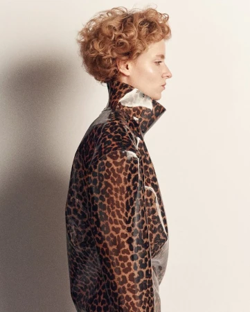 A woman with curly red hair wearing a shiny leopard-print jacket stands against a white backdrop, showcasing a stylish fashion portrait.