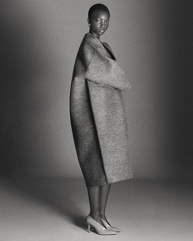 Studio shot of a Black woman in a grey coat and heels. Full-length portrait on a neutral backdrop. Fashion editorial style.