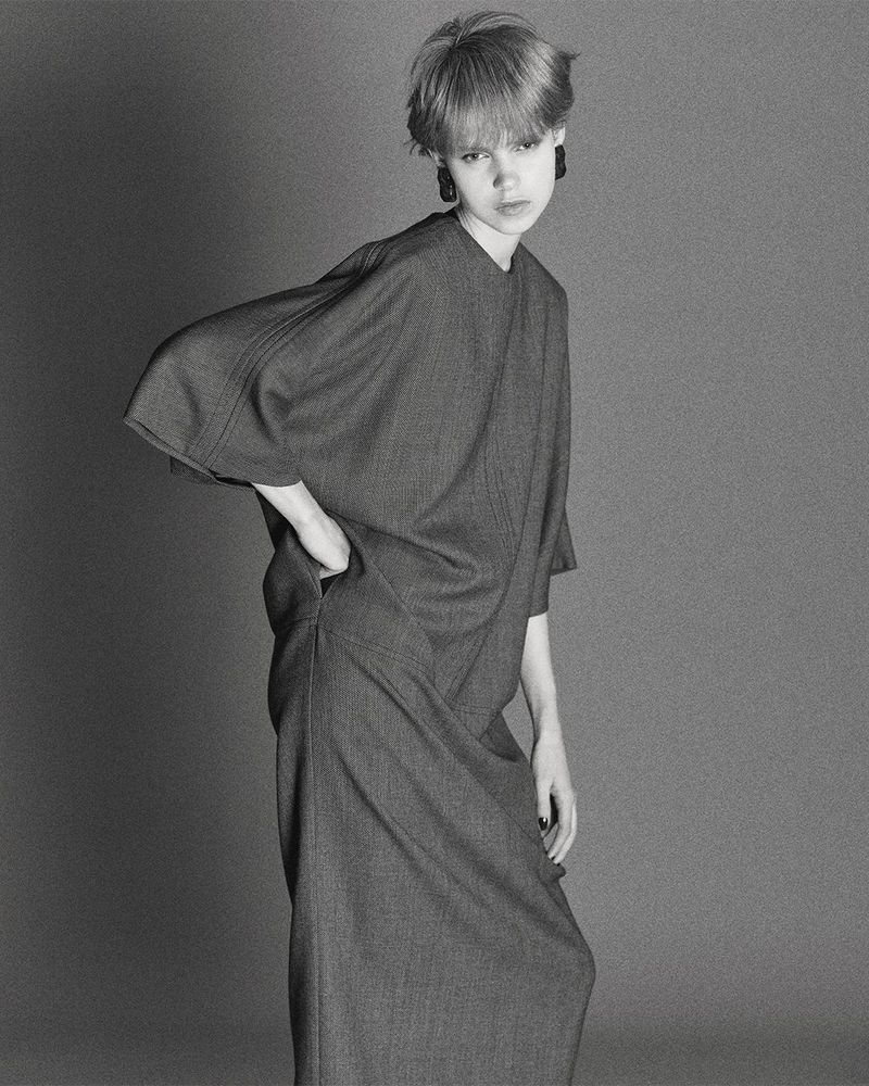 A woman with short blonde hair poses in a grey dress against a grey background in a studio shot, emphasizing fashion and design.