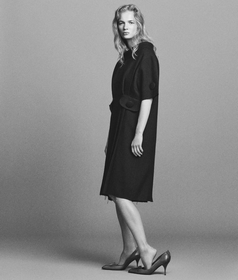 A blonde-haired model poses in a black dress with one foot on a pair of high heels against a gray backdrop in a studio fashion shot.