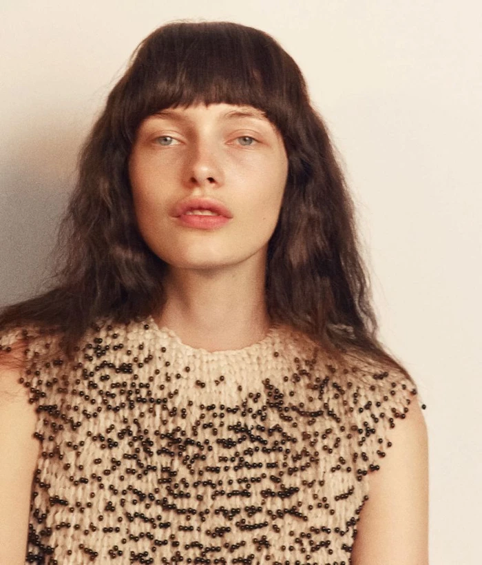 Portrait of a woman with dark hair and bangs wearing a cream top embellished with small dark beads against a plain background