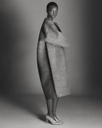 A Black model in an oversized sculptural grey wool coat and pointed-toe kitten heels poses in profile against a grey studio backdrop.