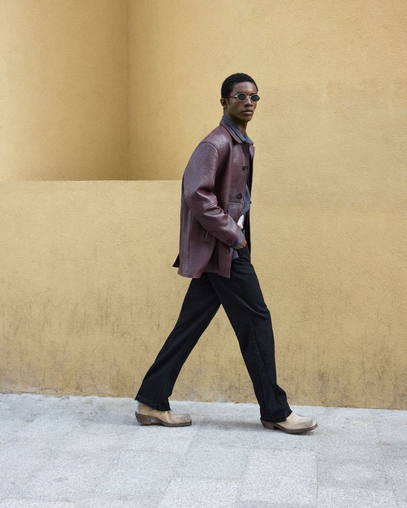 An adult Black man in a maroon jacket and dark jeans strides confidently against a warm-toned wall, showcasing casual style.
