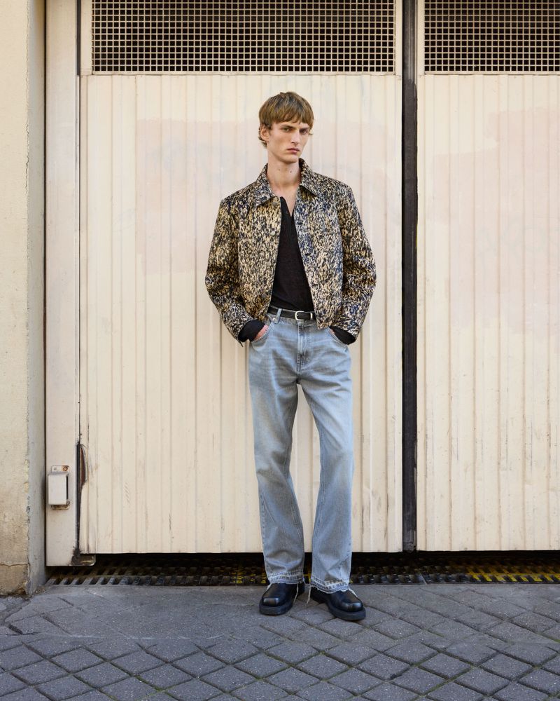 A blond male model stands in front of a door wearing a patterned jacket, black t-shirt, light wash jeans, and black shoes.