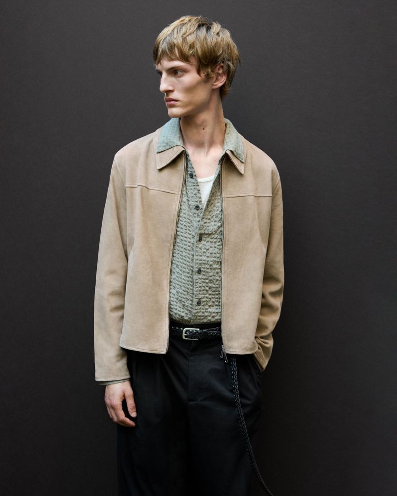 Profile of a young adult male in an open collared shirt and jacket against a dark gray background in a medium shot, indoor studio.