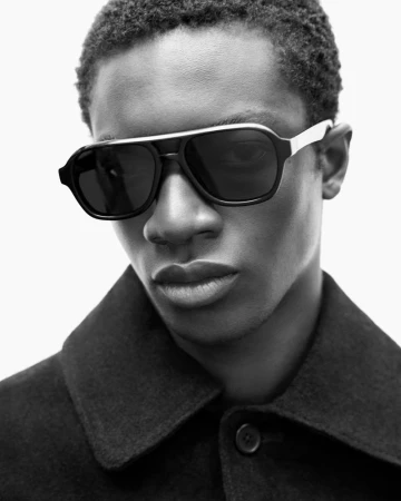 Close-up black and white portrait of a dark-skinned man wearing dark sunglasses and a wool coat against a bright white background.
