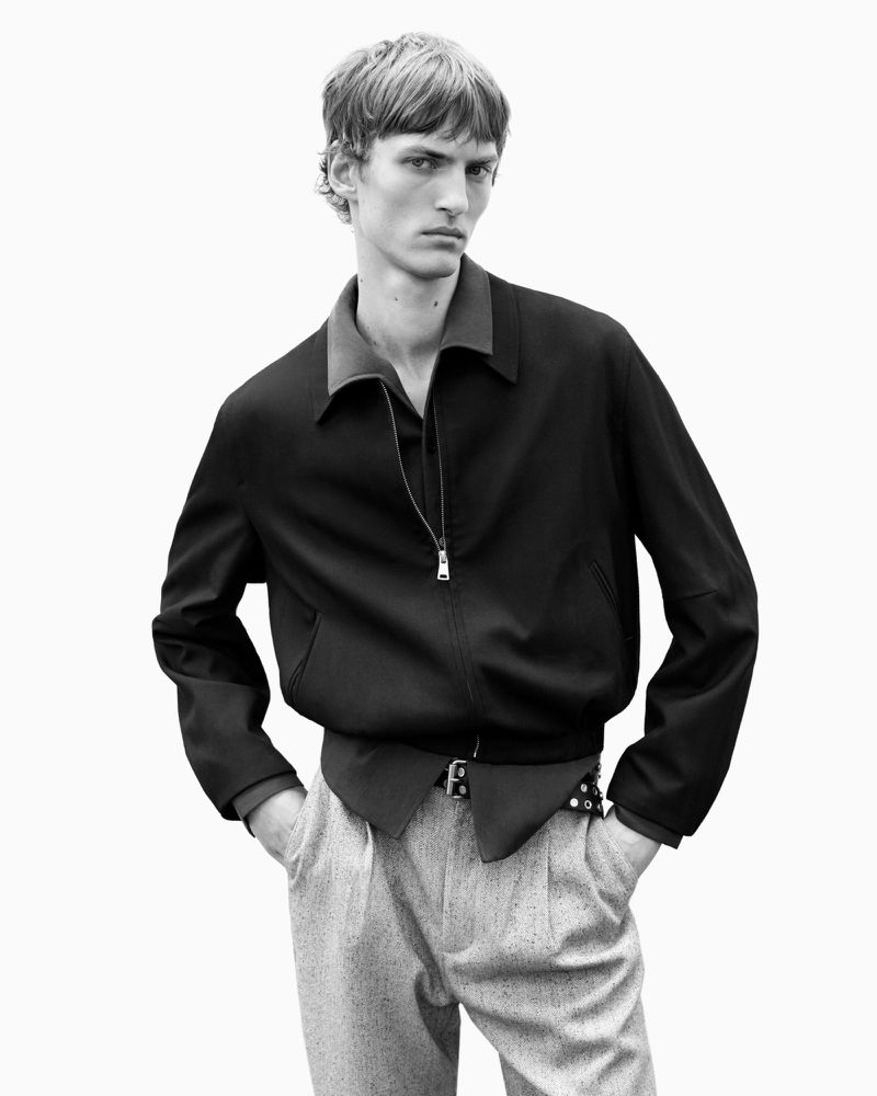 Monochrome portrait of a male model wearing a dark zipped jacket and light patterned trousers against a plain white background.