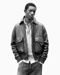 Black man wearing a leather jacket, cable knit sweater, and denim jeans against a white background