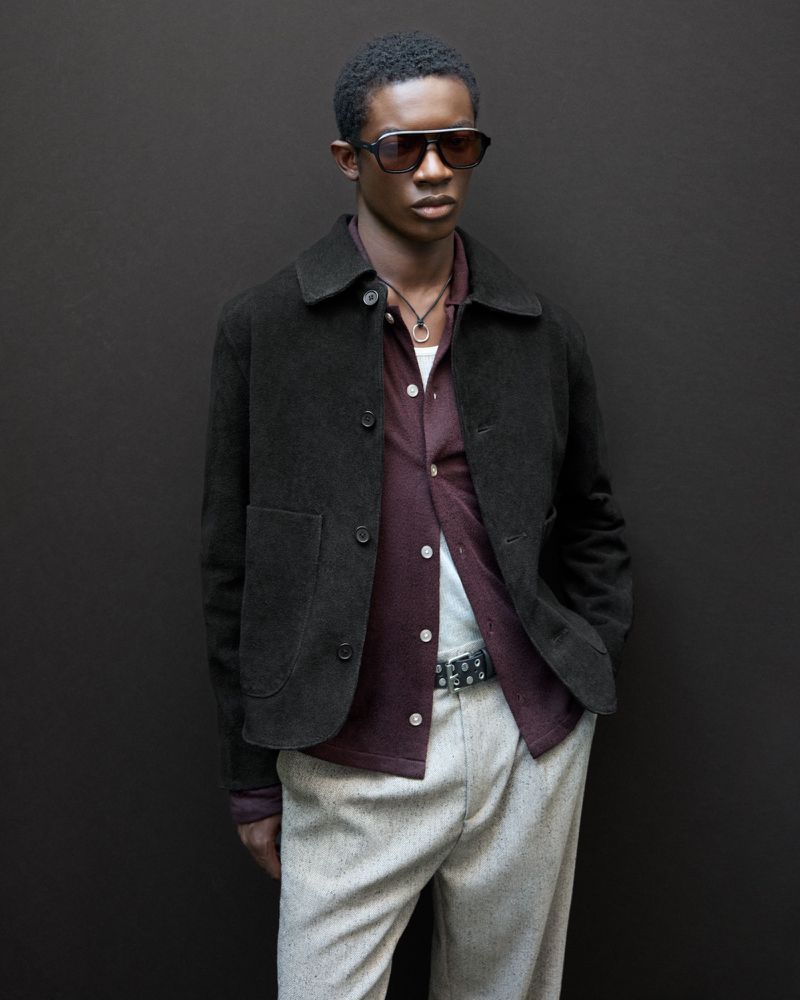 Model wearing dark sunglasses, brown suede jacket, burgundy shirt, and light grey trousers against dark background