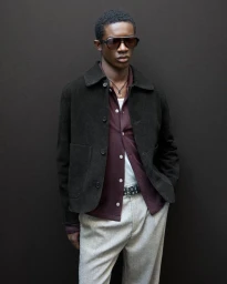 Model wearing dark sunglasses, brown suede jacket, burgundy shirt, and light grey trousers against dark background