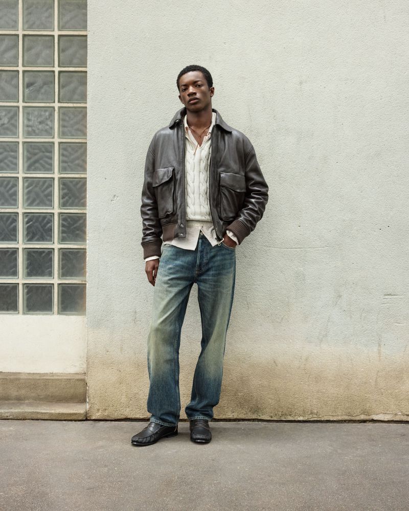 Model Feranmi Ajetomobi standing in a brown leather bomber jacket and relaxed blue jeans against a textured exterior concrete wall.