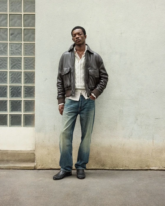 Model Feranmi Ajetomobi standing in a brown leather bomber jacket and relaxed blue jeans against a textured exterior concrete wall.