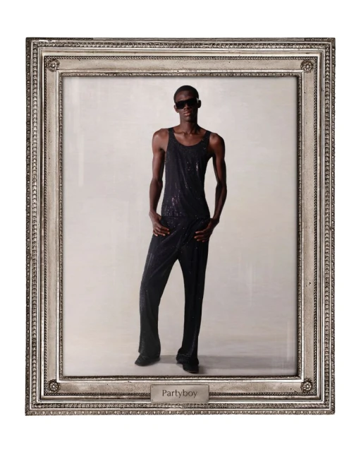 Person in black jumpsuit posing in silver picture frame against neutral background, illuminated by direct lighting and cool tones.