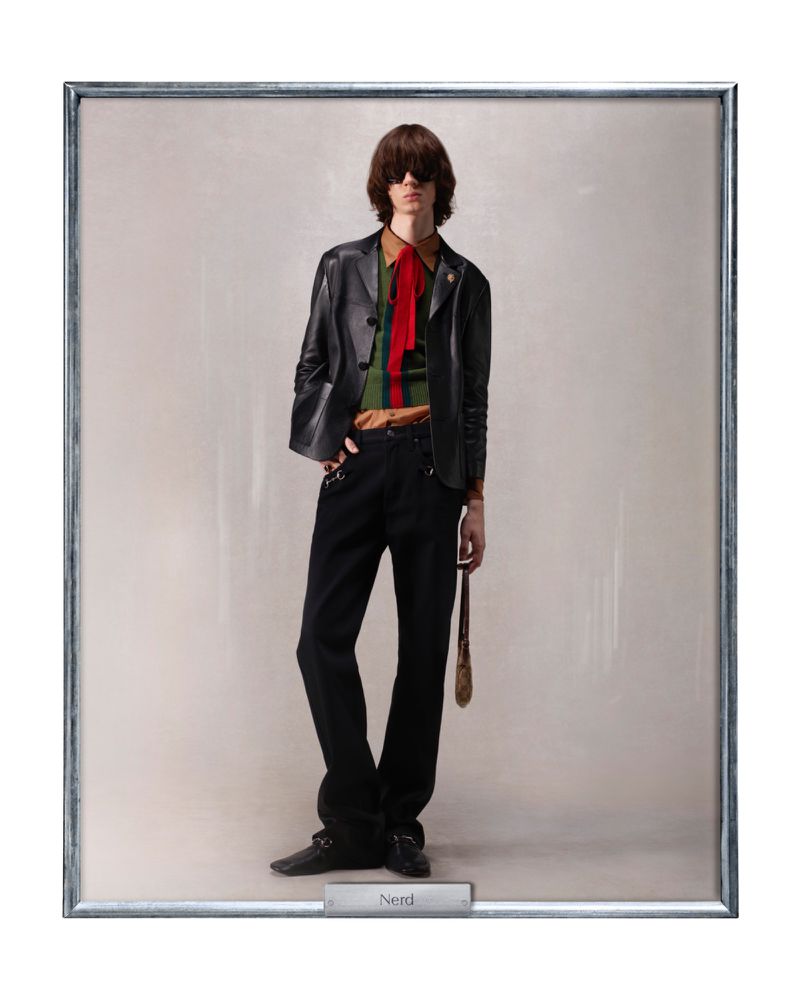 A man in a black leather jacket, green and brown striped shirt with a red tie stands in a silver frame, looking straight ahead.