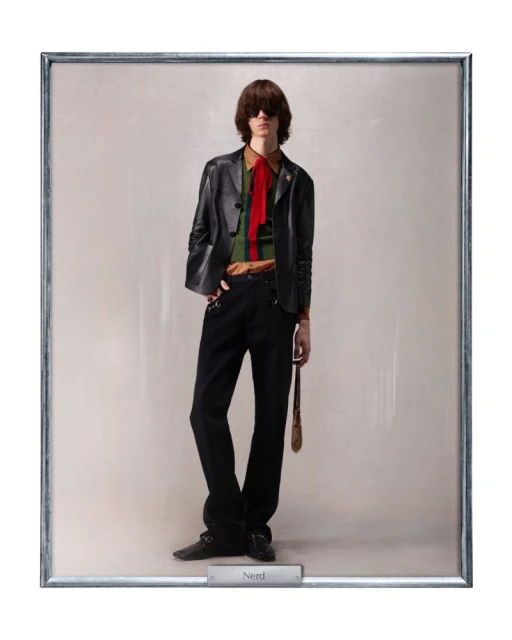 A man in a black leather jacket, green and brown striped shirt with a red tie stands in a silver frame, looking straight ahead.