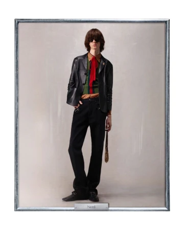 A man in a black leather jacket, green and brown striped shirt with a red tie stands in a silver frame, looking straight ahead.