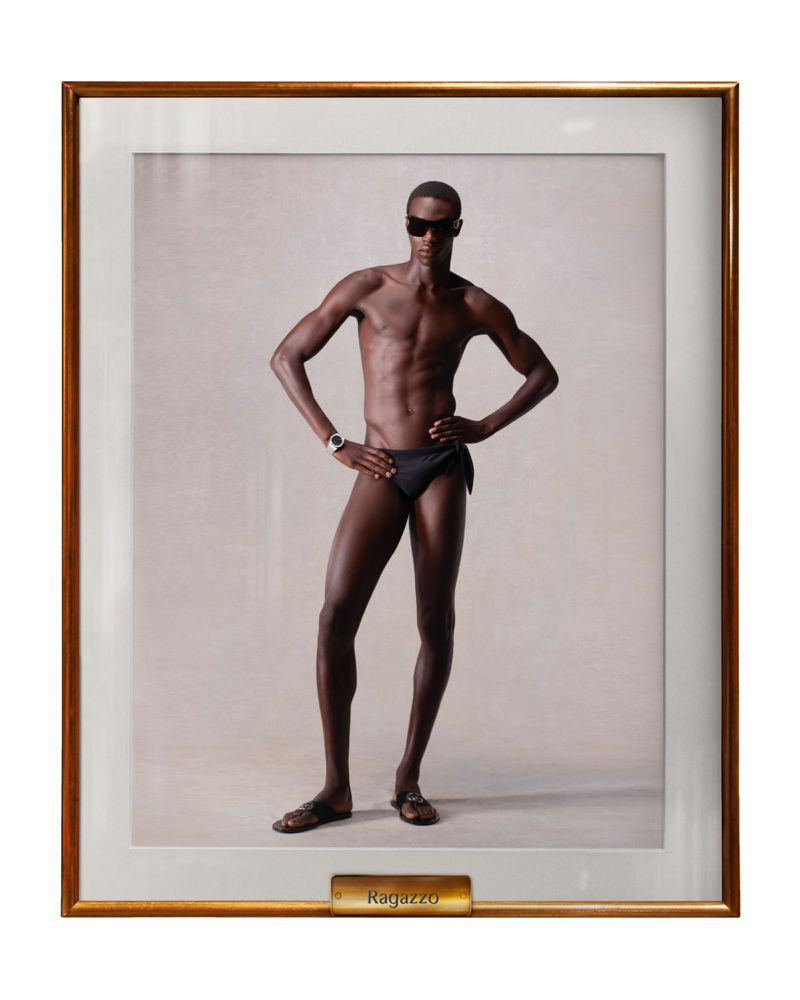 Framed studio portrait of a dark-skinned male model wearing a black swimsuit and sunglasses, posing with hands on hips in neutral light.