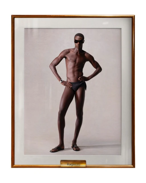 Framed studio portrait of a dark-skinned male model wearing a black swimsuit and sunglasses, posing with hands on hips in neutral light.