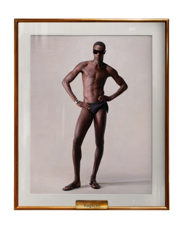 Framed studio portrait of a dark-skinned male model wearing a black swimsuit and sunglasses, posing with hands on hips in neutral light.