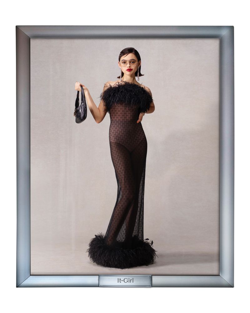 Elegant woman poses in a black sheer dress with feather trim, holding a black purse, set against a neutral background inside a picture frame.